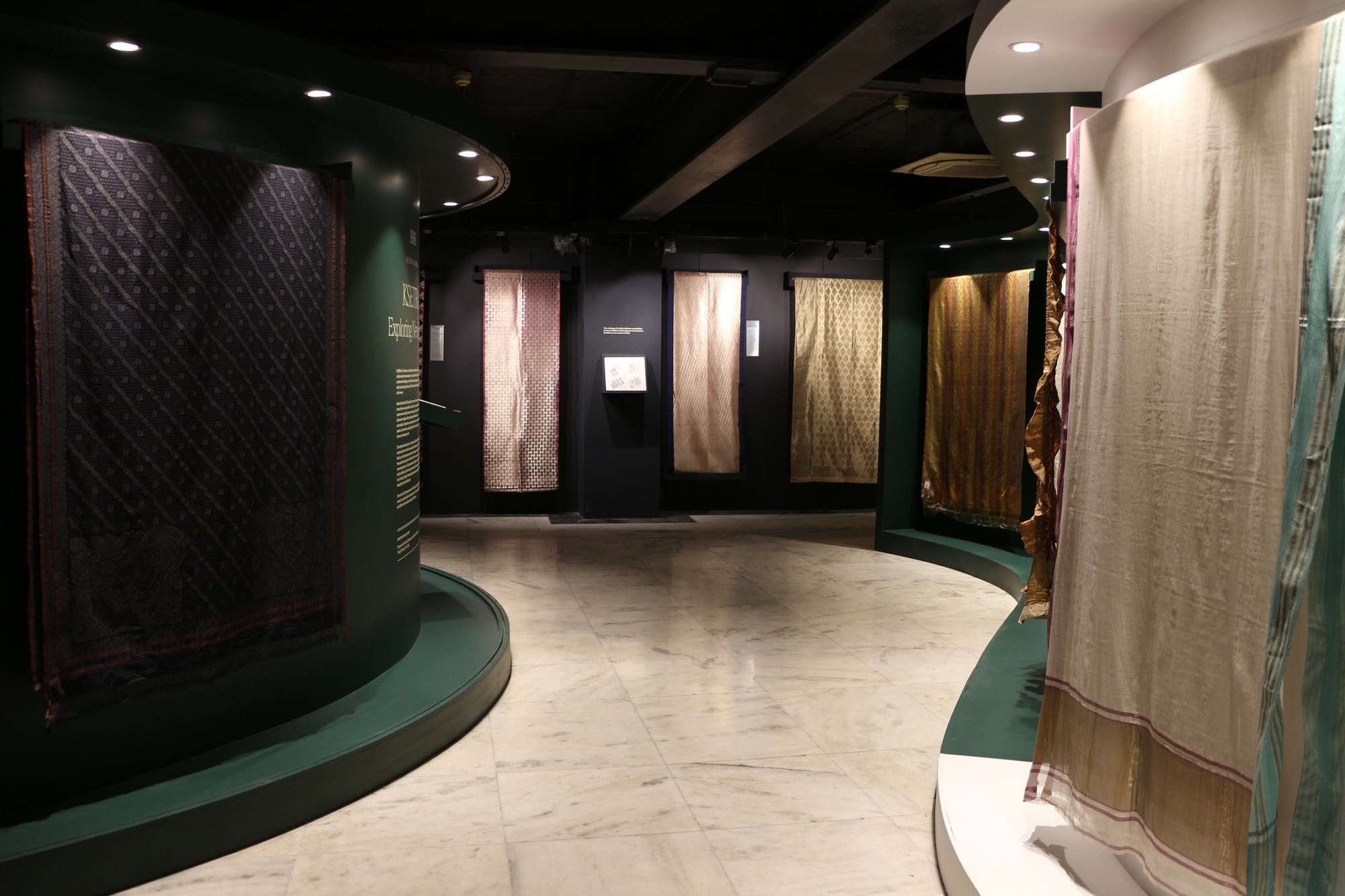 A curved gallery corridor at the Vishwakarma Gallery, Handloom Haat, Janpath: hand-woven textiles lit on dark green and charcoal display walls above a polished stone floor.